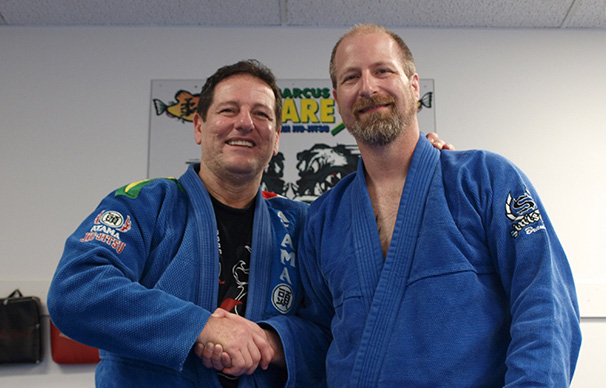 Two men are shaking hands and smiling. The man on the right, Marcus Soares, has an arm around the other man's shoulders. The man on the left, Bryan Rumble, is partially bald with facial hair.