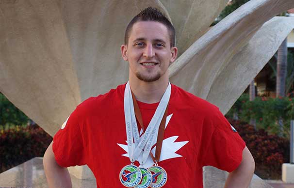 A man stands in front of a statue, he has a smile on his face and two silver medals and a bronze one around his neck.
