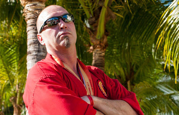 Darrell Warren, a bald man with facial hair and sunglasses stands with his arms crossed in front of palm trees. He looks serious but seems to be fighting a smile.