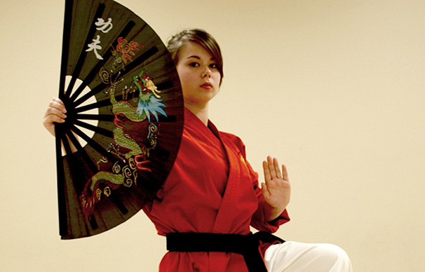 A young woman in her early 20s has a serious expression as she poses with a decorative fan. Her hair is pulled back and her left hand is raised palm out as her left hand holds the fan.