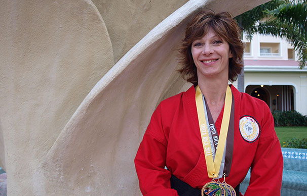 A woman with short cropped hair is smiling. She has a gold, silver and bronze medal around her neck. There are palm trees in the background.